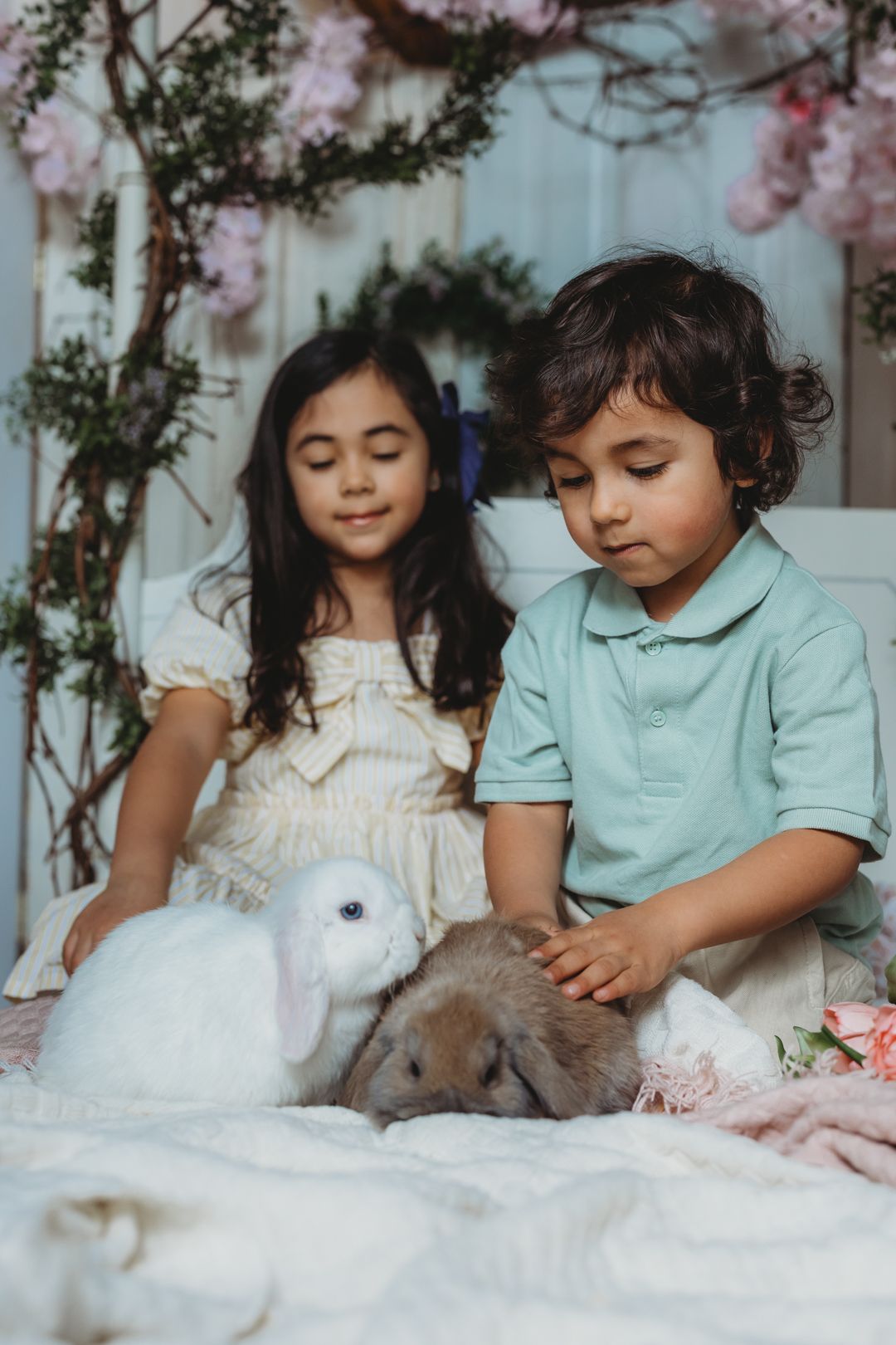 Two young siblings gently petting white and brown rabbits on bed with floral decorations.