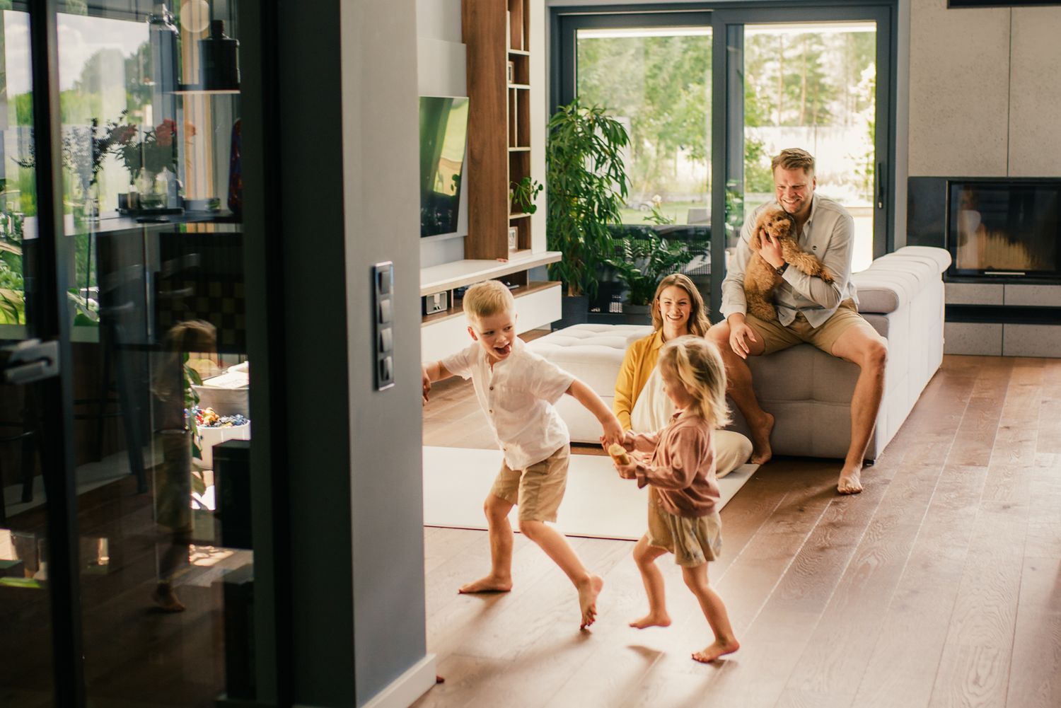 Children dancing barefoot in modern living room while father watches from couch smiling.