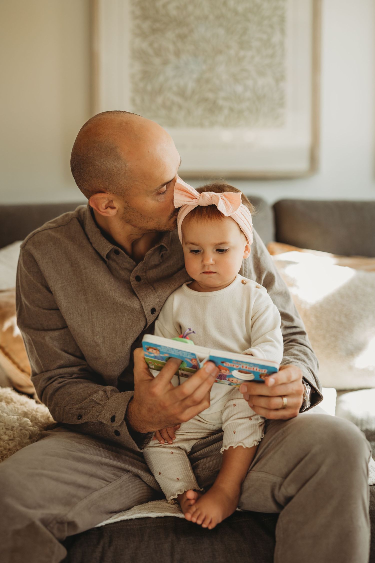 Father kissing baby girl wearing pink bow headband while reading a colorful book together on a cozy sofa.