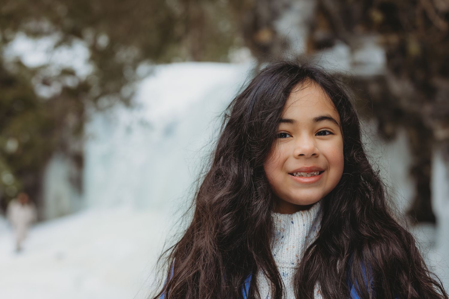 Smiling young girl with long dark hair posing in snowy winter forest background.