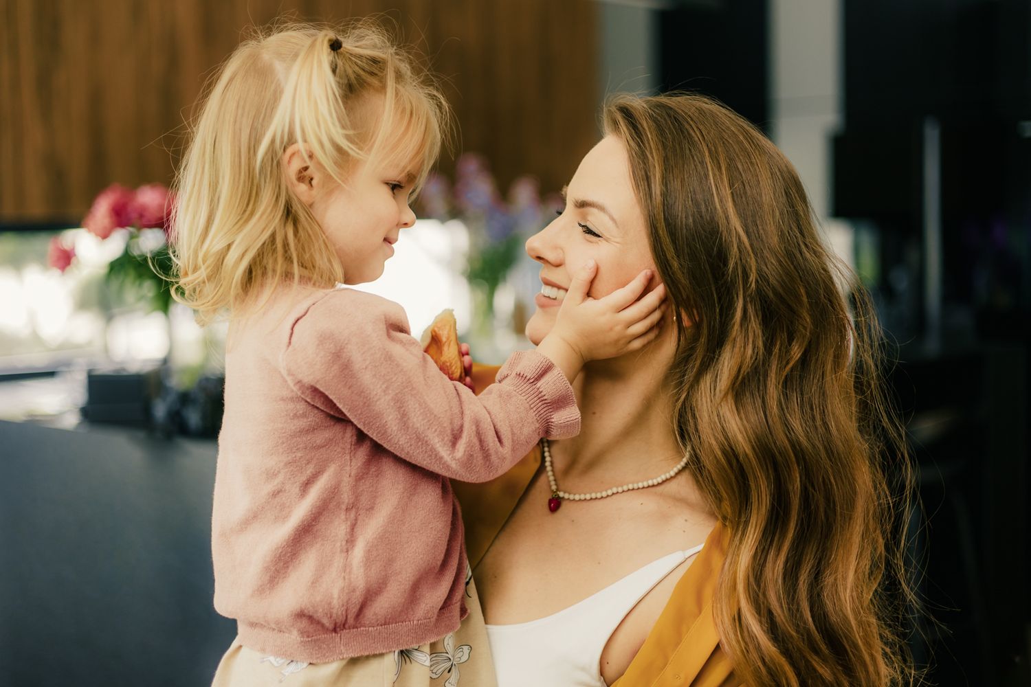 A sweet toddler girl in pink gently touches her smiling mothers face in a warm indoor setting.