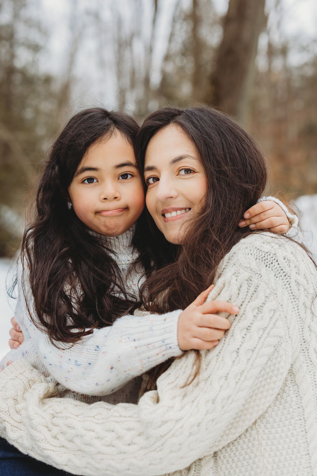 A mother and daughter in white sweaters share a warm embrace outdoors in a snowy winter forest.