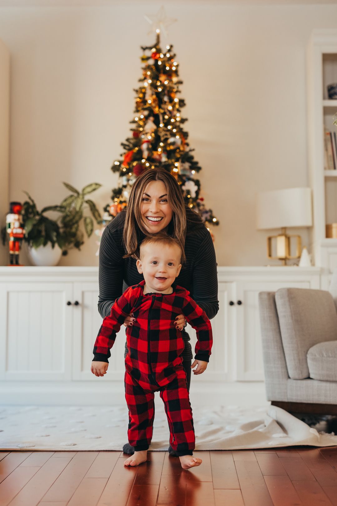 A smiling woman helps her baby in red plaid pajamas stand in front of a decorated Christmas tree indoors.