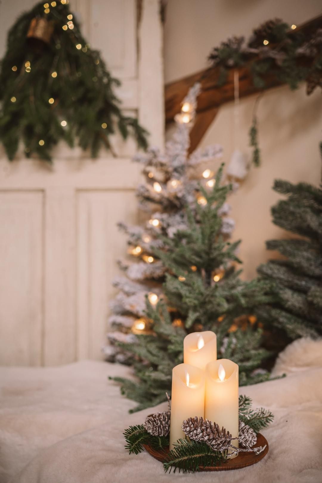 Three white candles on a wooden tray with pine cones, surrounded by Christmas trees and warm fairy lights.