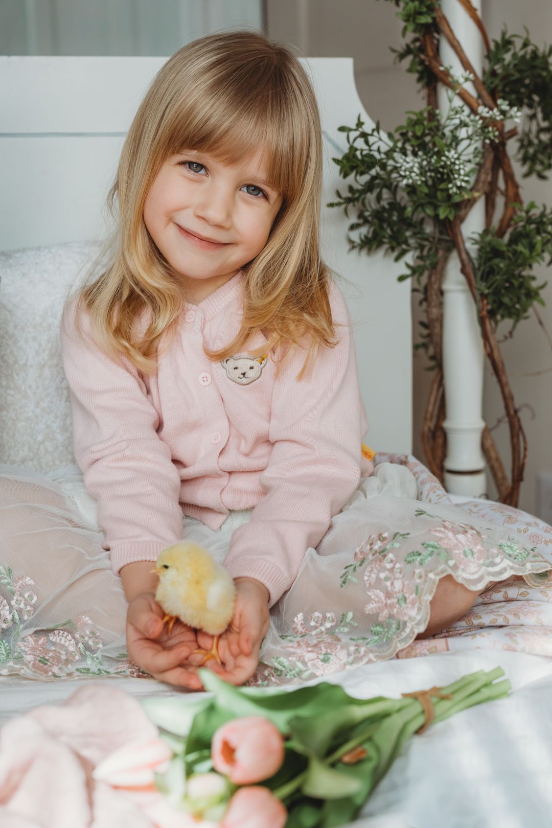 Smiling blonde girl in pink outfit sitting on bed holding a yellow baby chick with tulips nearby.