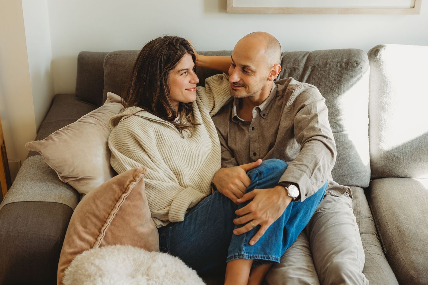 Couple sitting together on a cozy gray sofa, smiling and looking at each other in a warm living room.