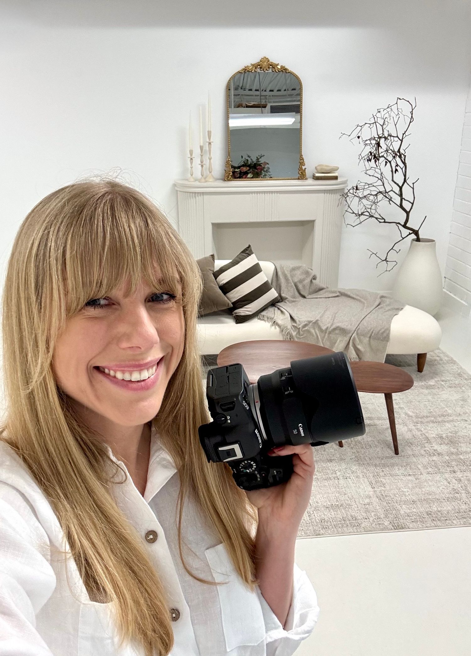 Smiling blonde woman holding a camera in a bright, modern living room with white decor and fireplace.