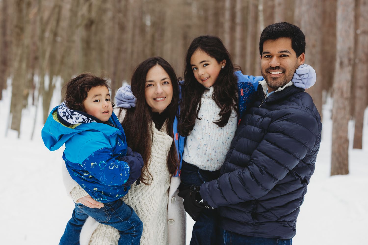 Family of four smiling in snowy winter forest, parents holding children in warm colorful jackets.