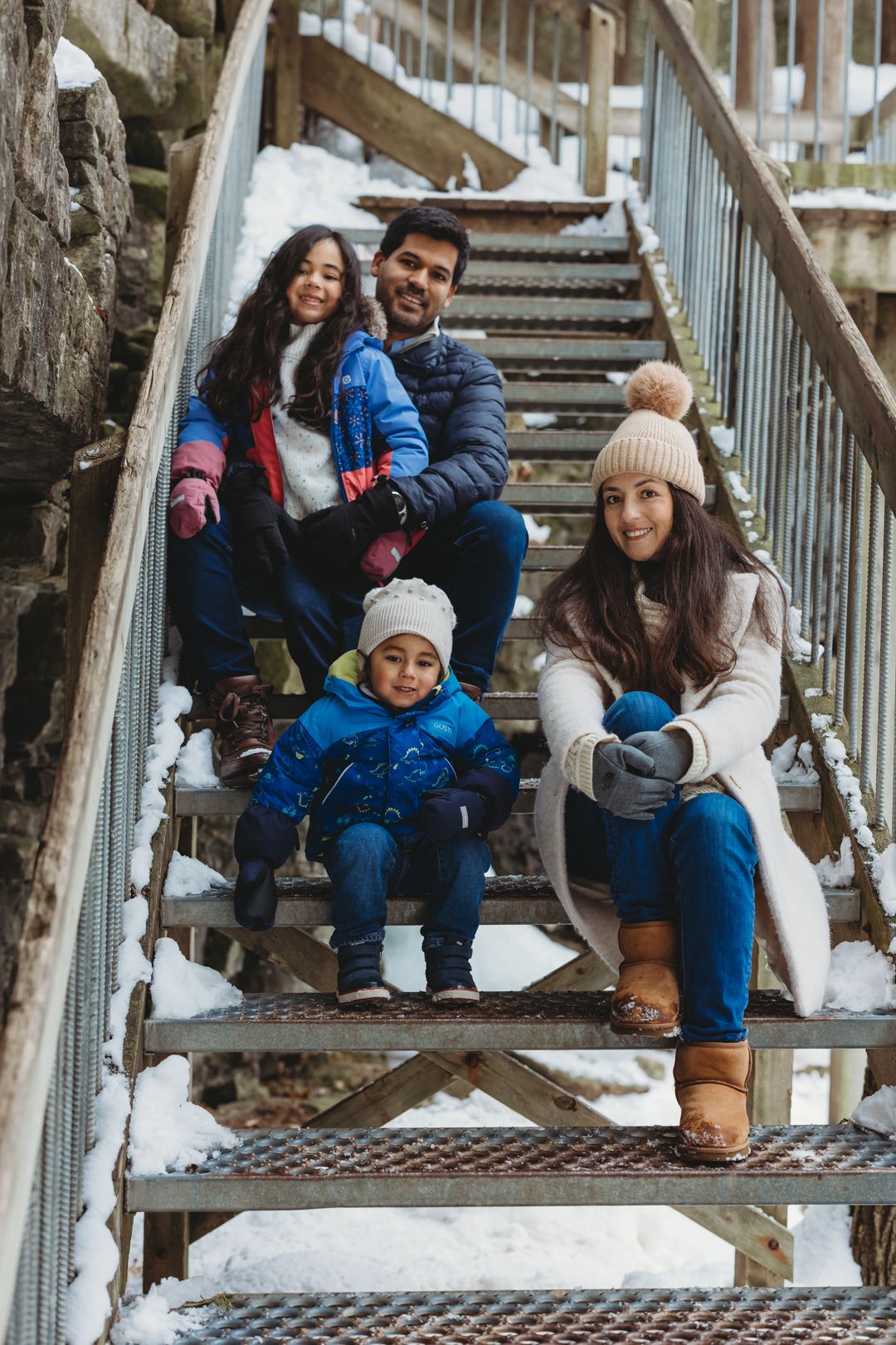 Happy family of four sits together on snowy metal stairs during a winter outing.