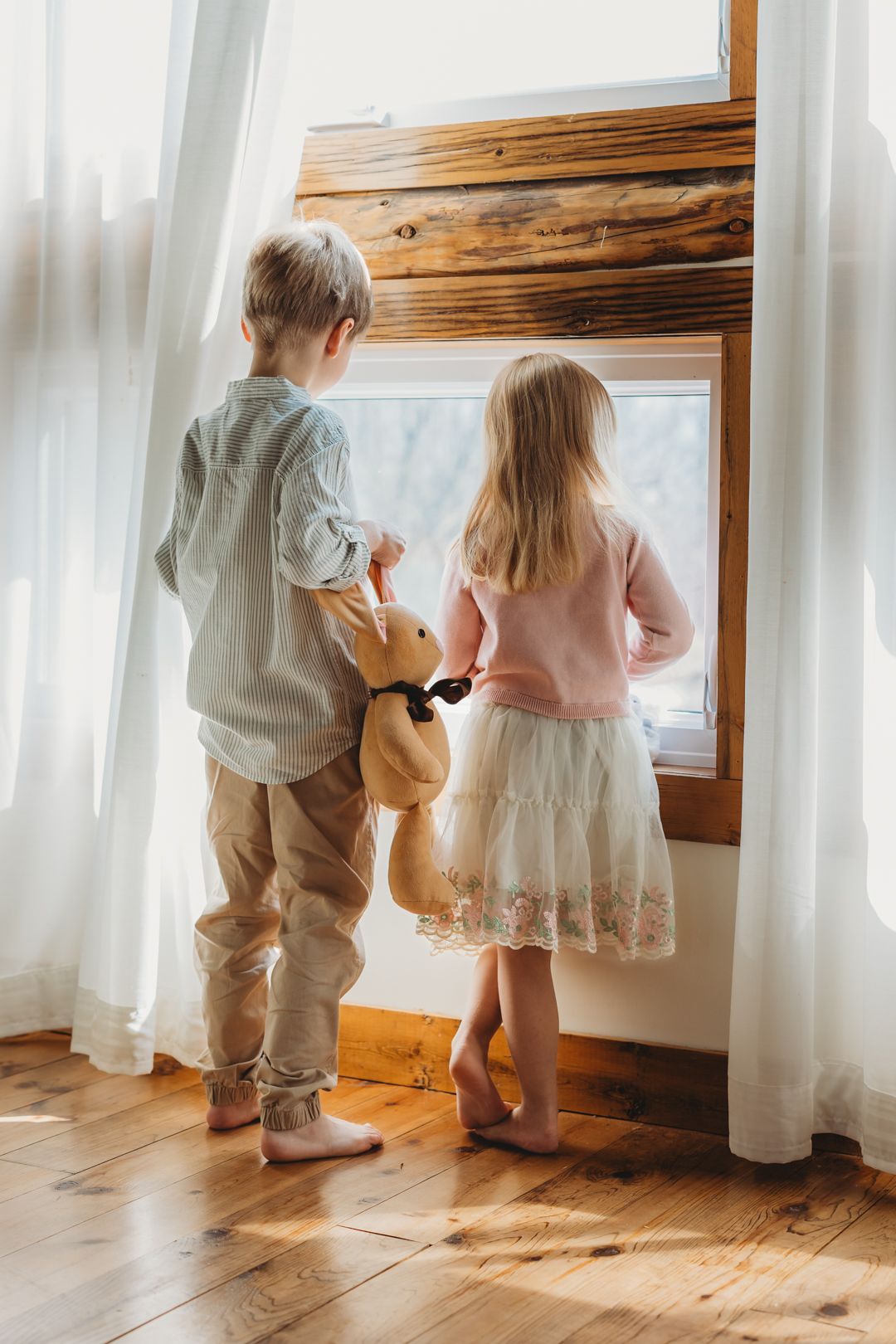 Two young children stand at a sunlit window, boy holding teddy bear, girl in pink sweater, on hardwood floors.