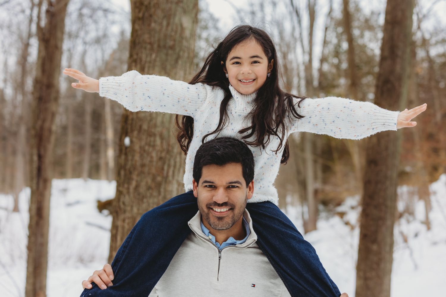 Happy girl with arms spread wide sitting on fathers shoulders in snowy winter forest.