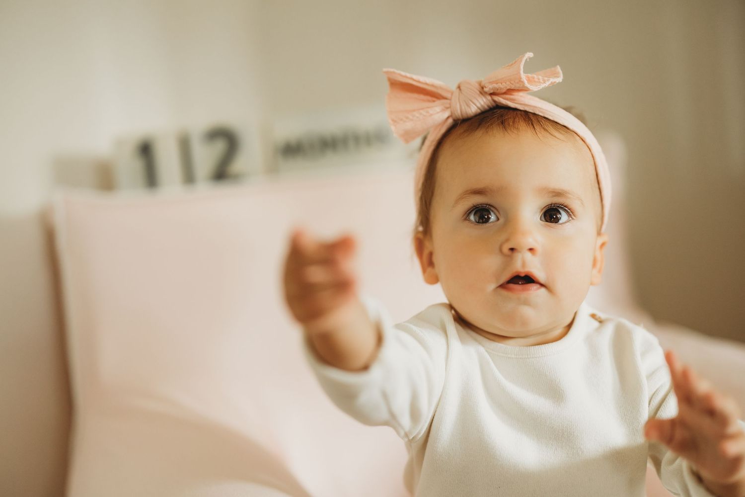 Adorable baby girl in white onesie and pink bow headband reaching out her hands, sitting in a nursery.