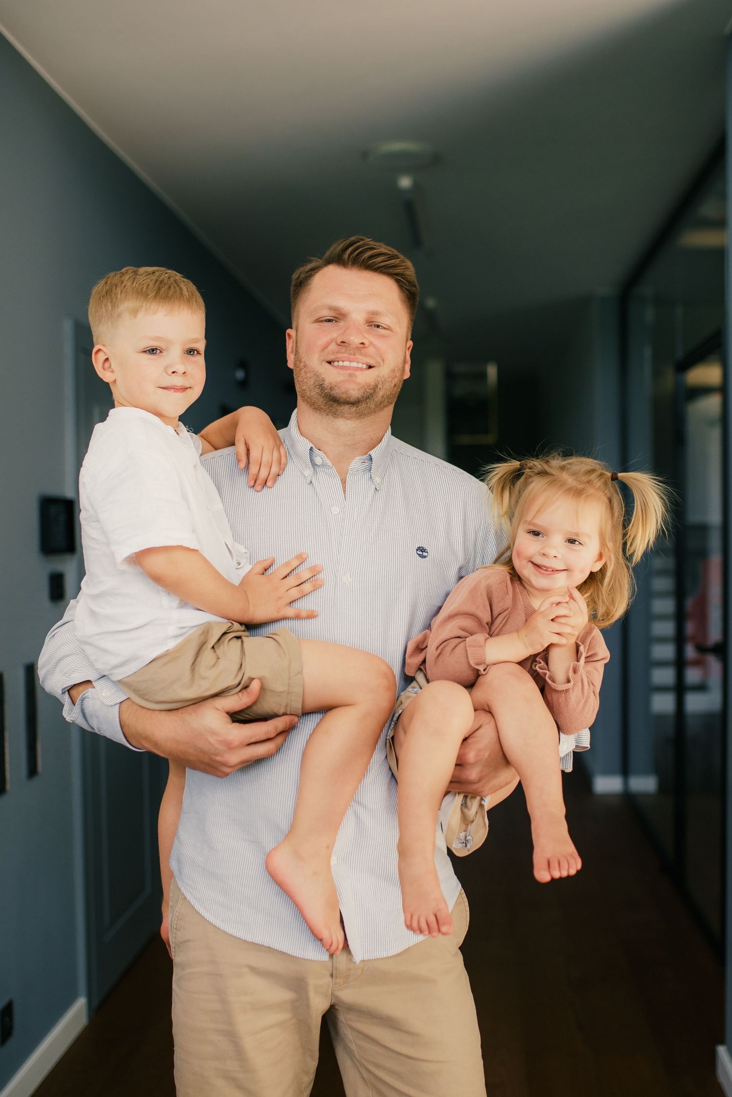 Smiling father holding his young son and toddler daughter in a hallway.