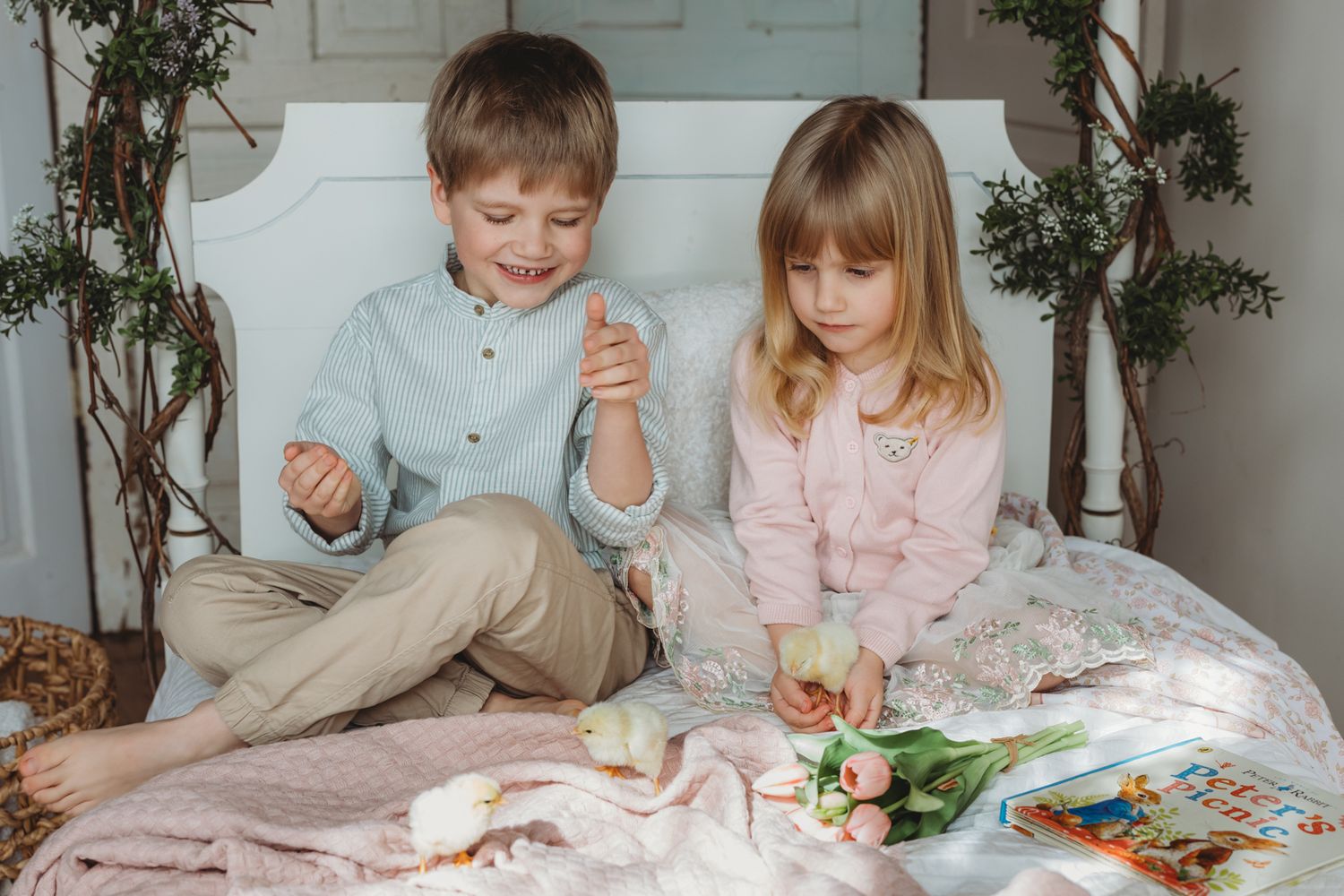 Two young children sitting on a bed reading a book together surrounded by tulips and spring decorations.