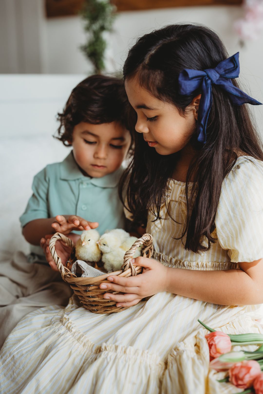 Two children examine baby chicks in a wicker basket, girl in yellow dress with blue bow, boy in mint shirt.