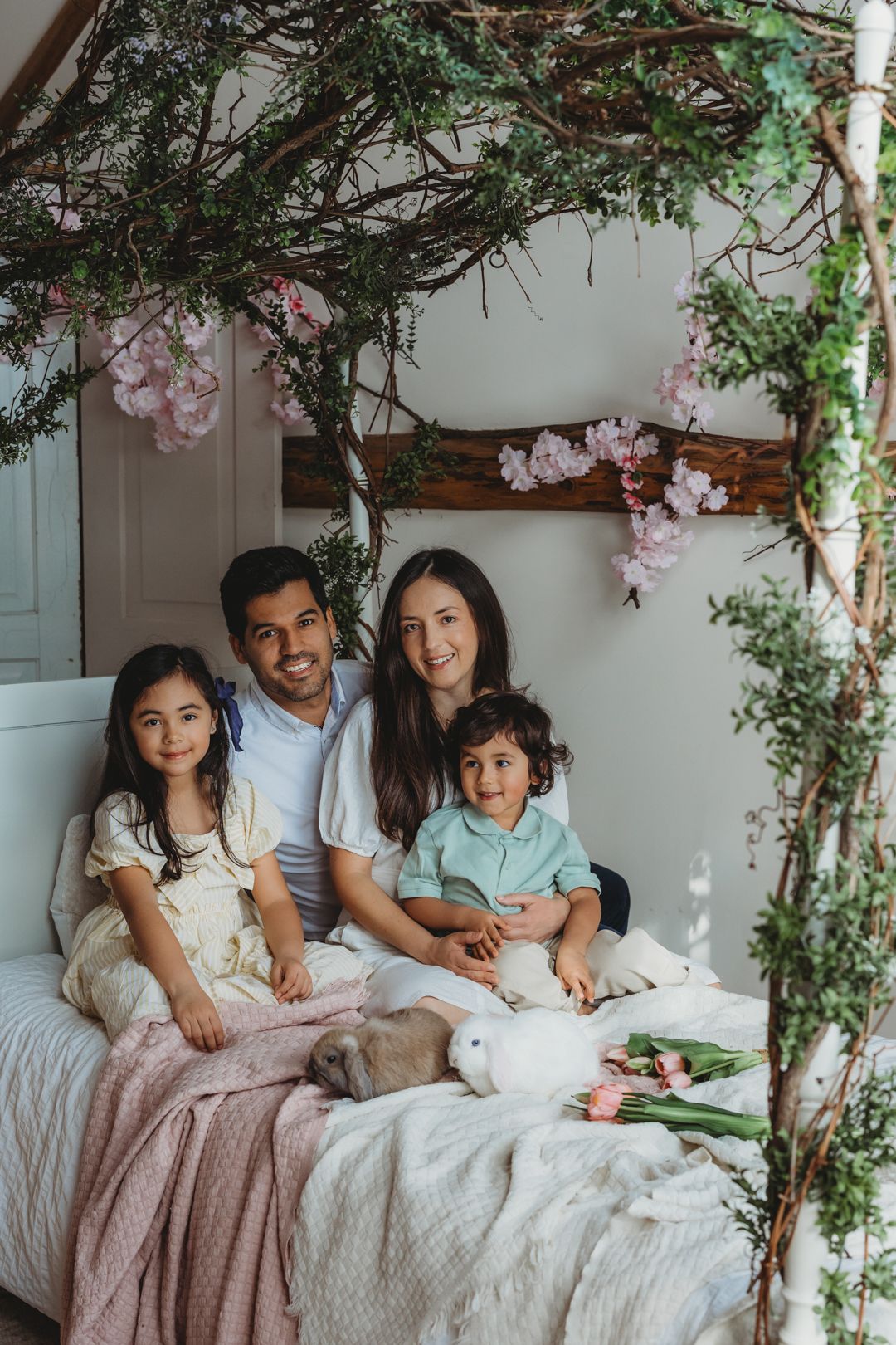 Happy family of four sits on white bed under floral arch with rabbits and spring tulips.