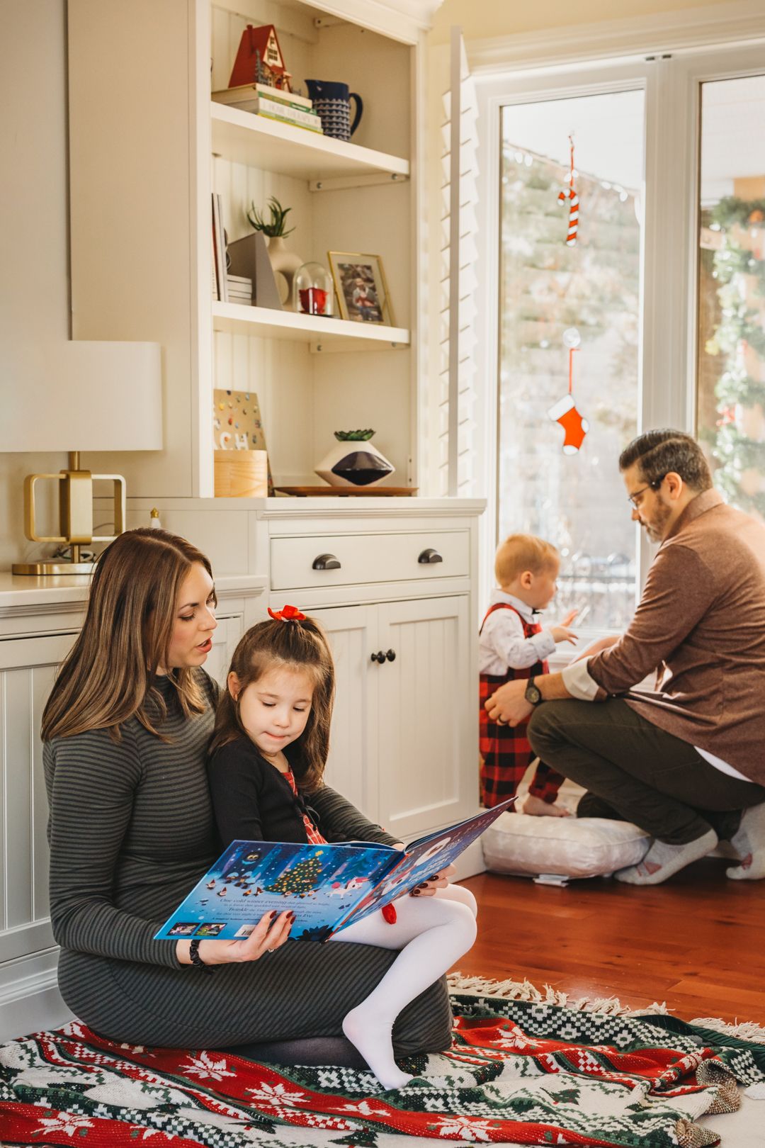 Family reading a Christmas book together on a holiday rug, with festive decorations and a snowy window in the background.