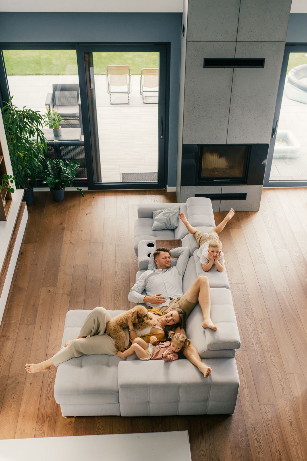 Top view of family with dog resting on gray sectional sofa in modern living room with fireplace.