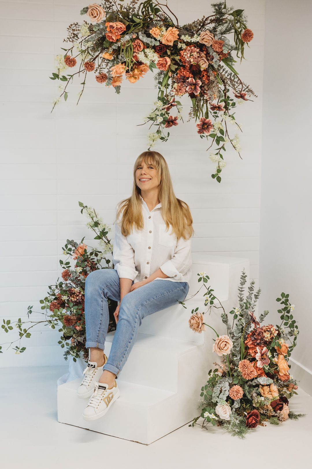 Happy blonde woman in casual outfit seated on white stairs with beautiful orange and red flower decor.