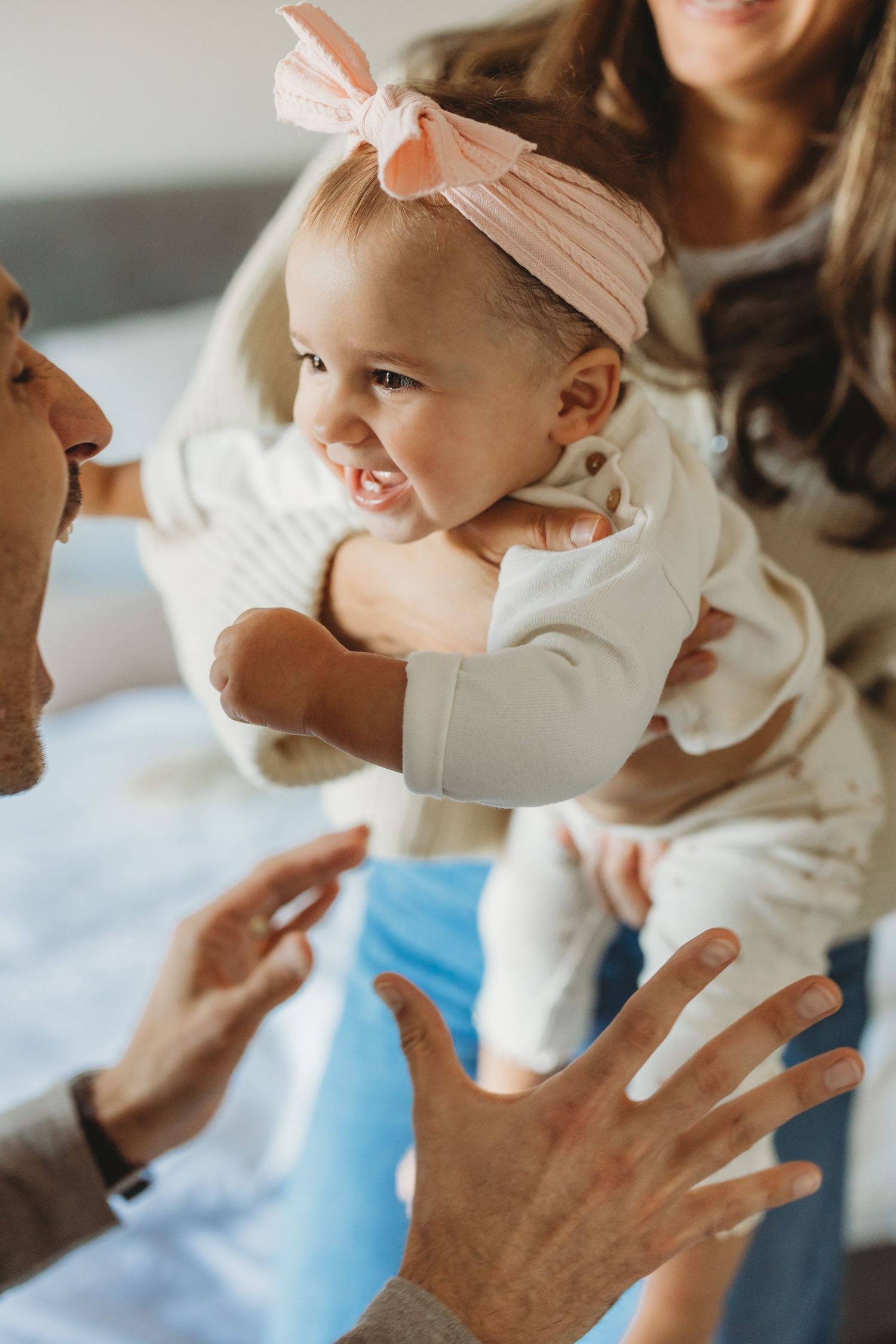 Parents playing with their smiling baby girl wearing a pink bow headband in a bright home setting.