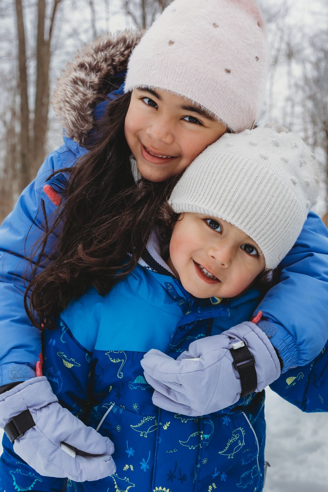 Two young siblings hugging and smiling in snowy winter setting, wearing blue jackets and knit hats.