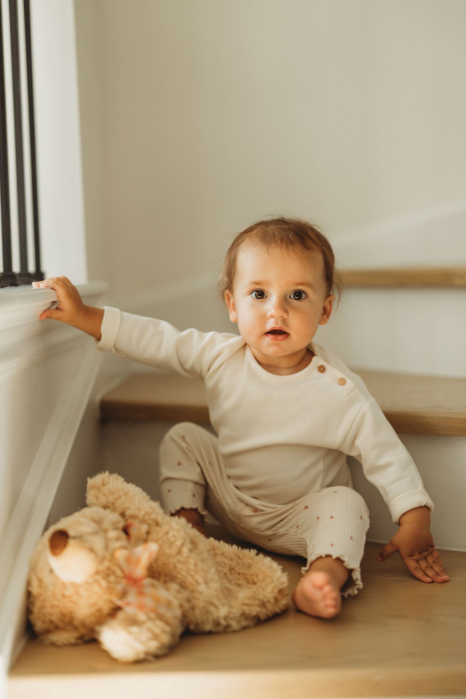 Cute baby in cream outfit sitting on wooden stairs with a teddy bear toy.