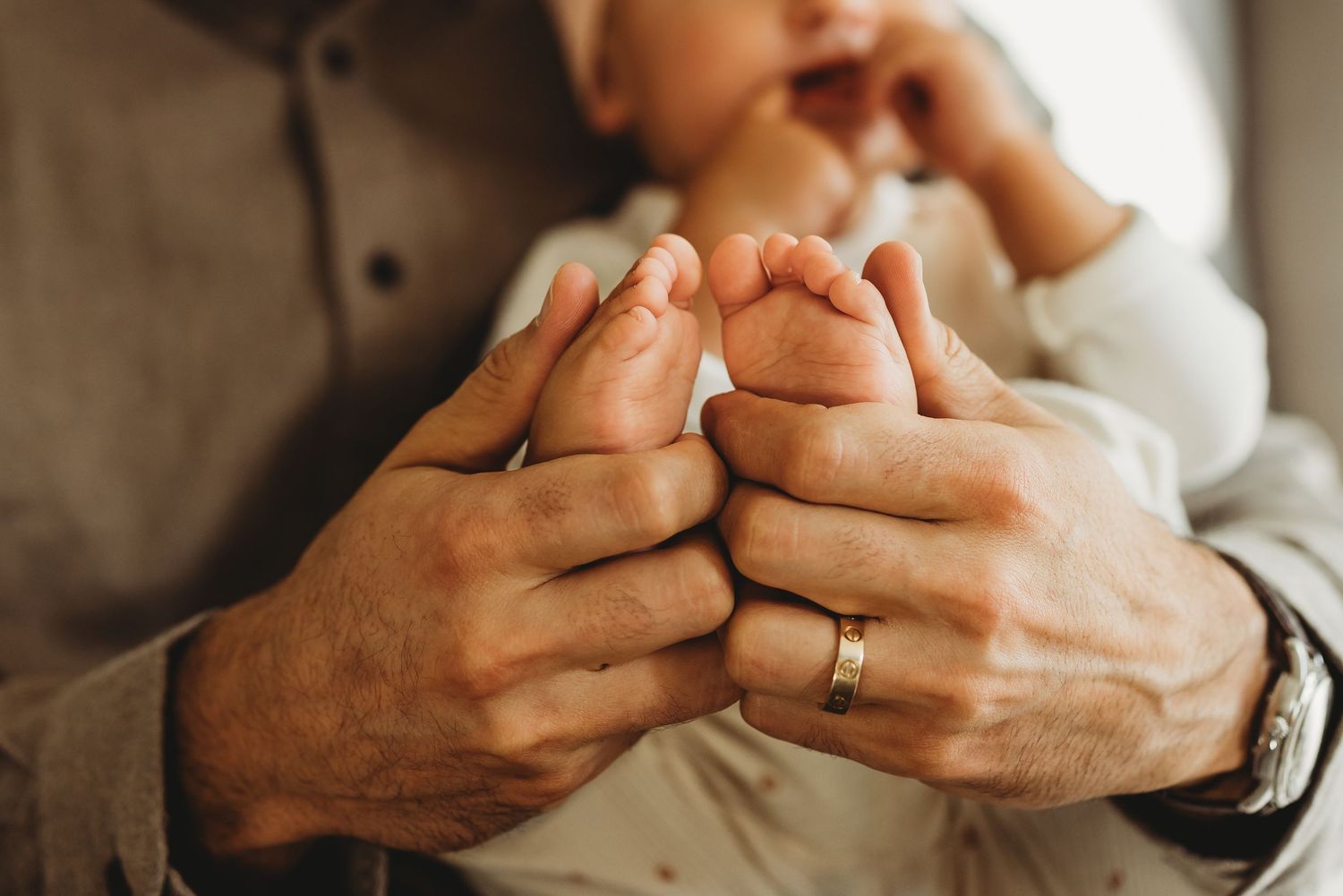 Close-up of a father's hands cradling tiny newborn baby feet, with a gold wedding ring visible.