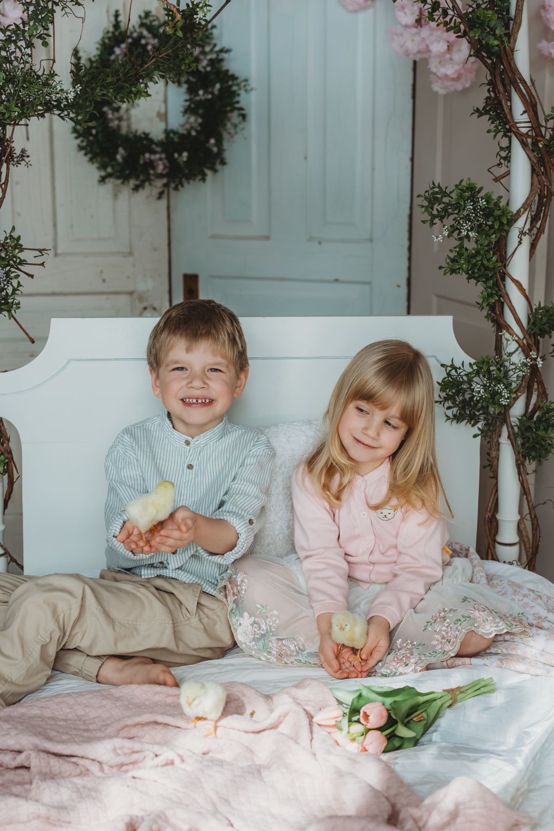 Two smiling siblings sit on a bed holding baby chicks surrounded by spring floral Easter decorations.