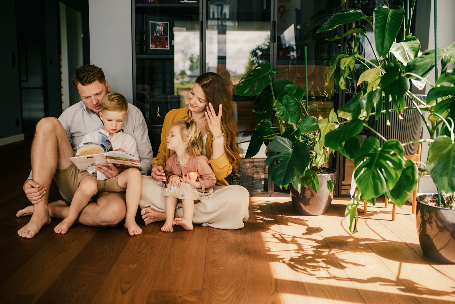A happy family of four sits on a sunny hardwood floor reading a book surrounded by lush indoor plants.