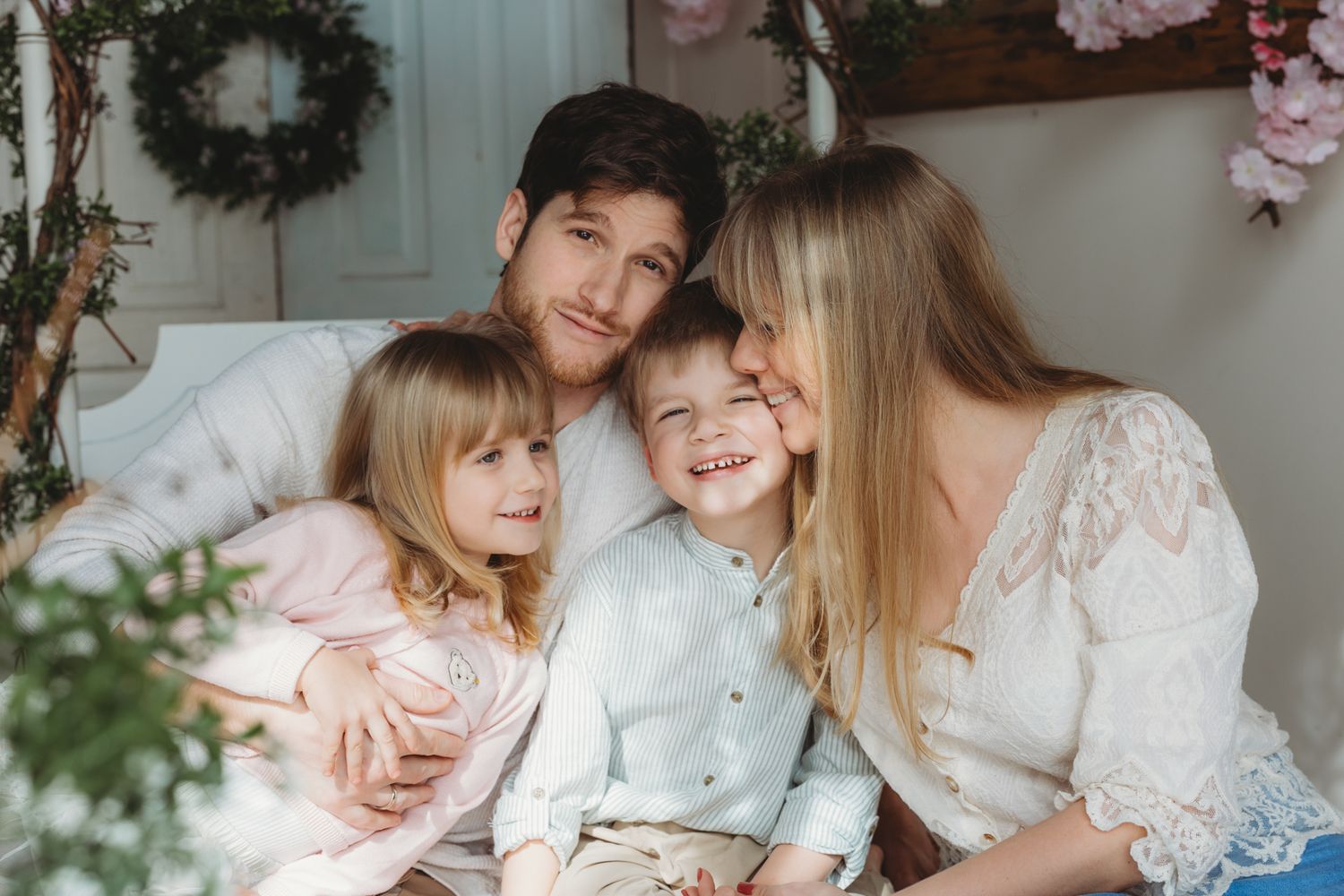 Happy family of four posing together on a bed with Christmas wreaths and pink floral decorations in background.