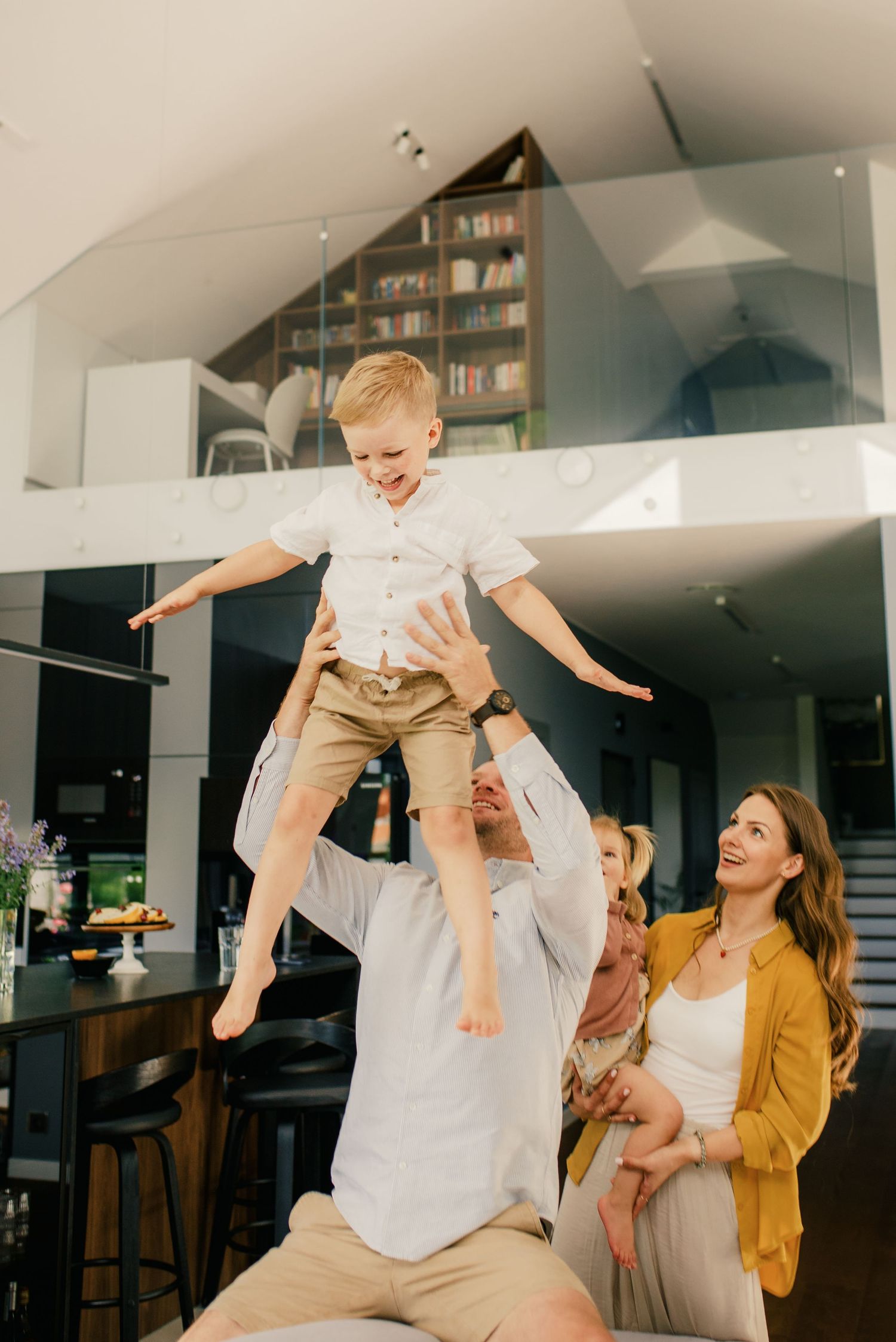 Laughing father lifting excited young boy in air while mother watches joyfully in modern home interior.