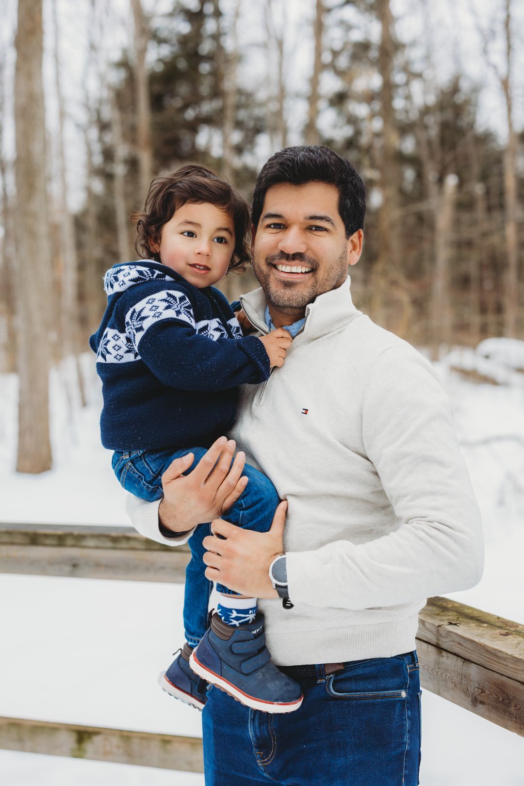 A smiling father in a white Tommy Hilfiger pullover holds his toddler son in a snowy winter park.