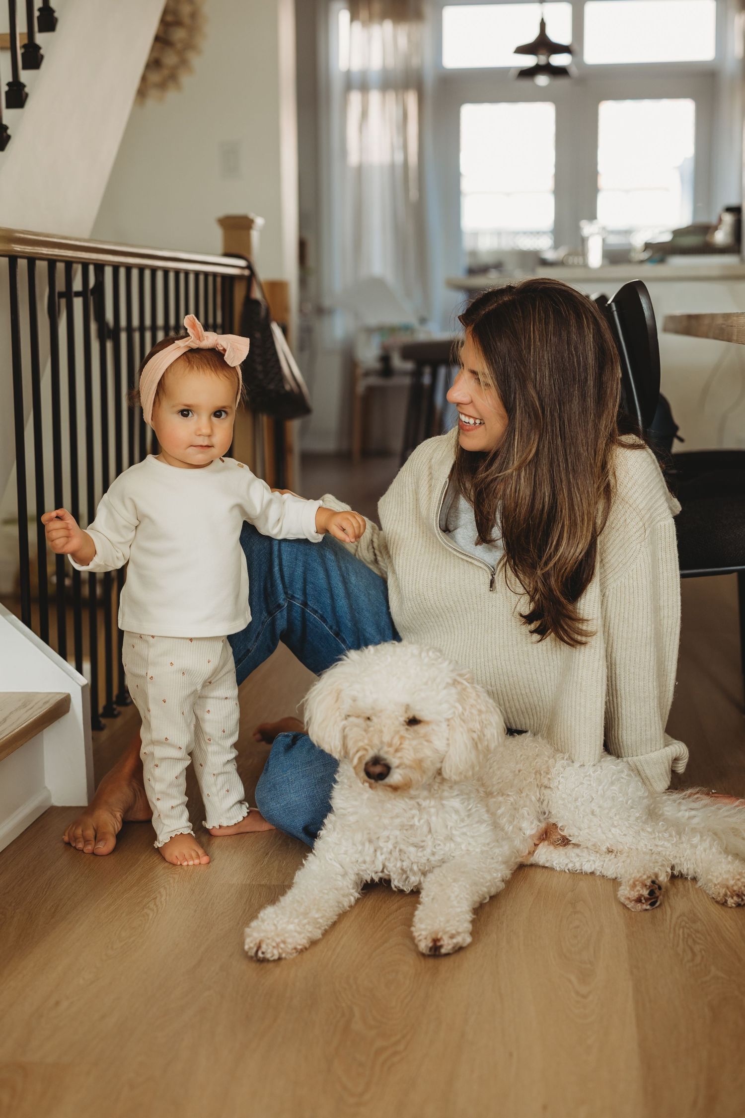 A mother sits on hardwood floor smiling at her baby girl standing nearby, with a white fluffy dog resting beside them.
