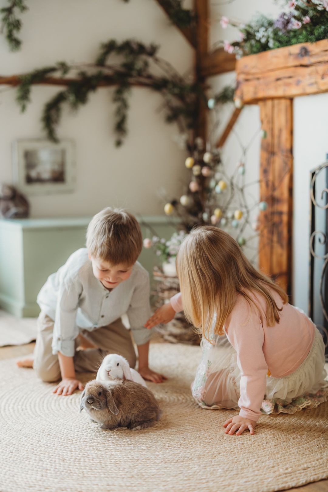 Young boy and girl kneel on rug petting a brown rabbit near Easter decorations and fireplace.