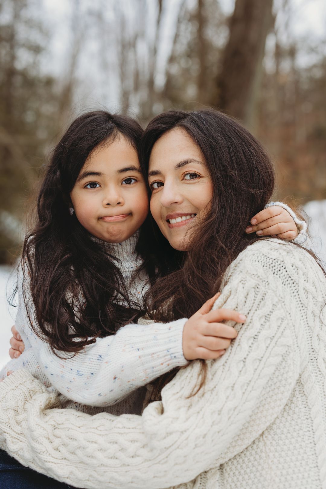A mother and daughter in white knit sweaters share a warm embrace outdoors in a snowy winter forest.