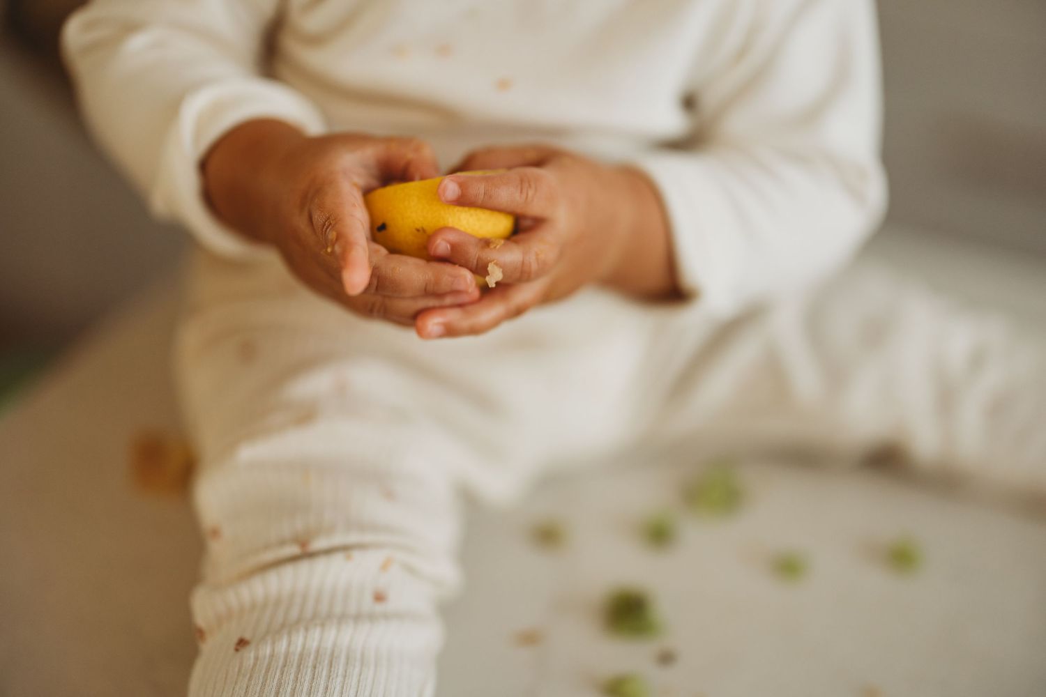 Child in white outfit holding a yellow lemon with scattered green pieces on a light surface.
