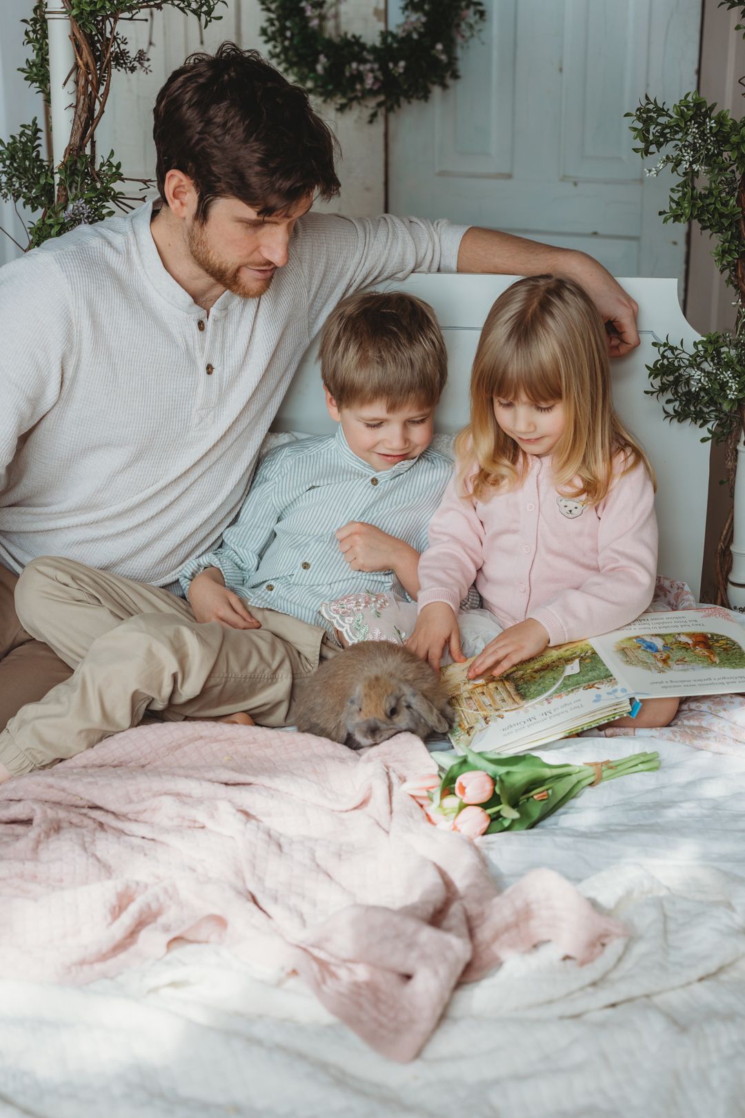 Father reads a storybook with two young children and a puppy on a cozy bed with tulips.