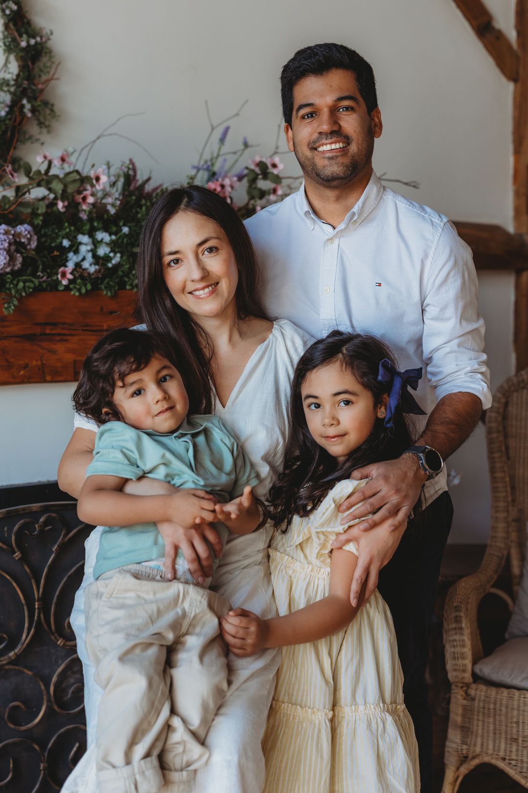 Happy family of four posing together with floral backdrop, parents holding young children in bright spring setting.