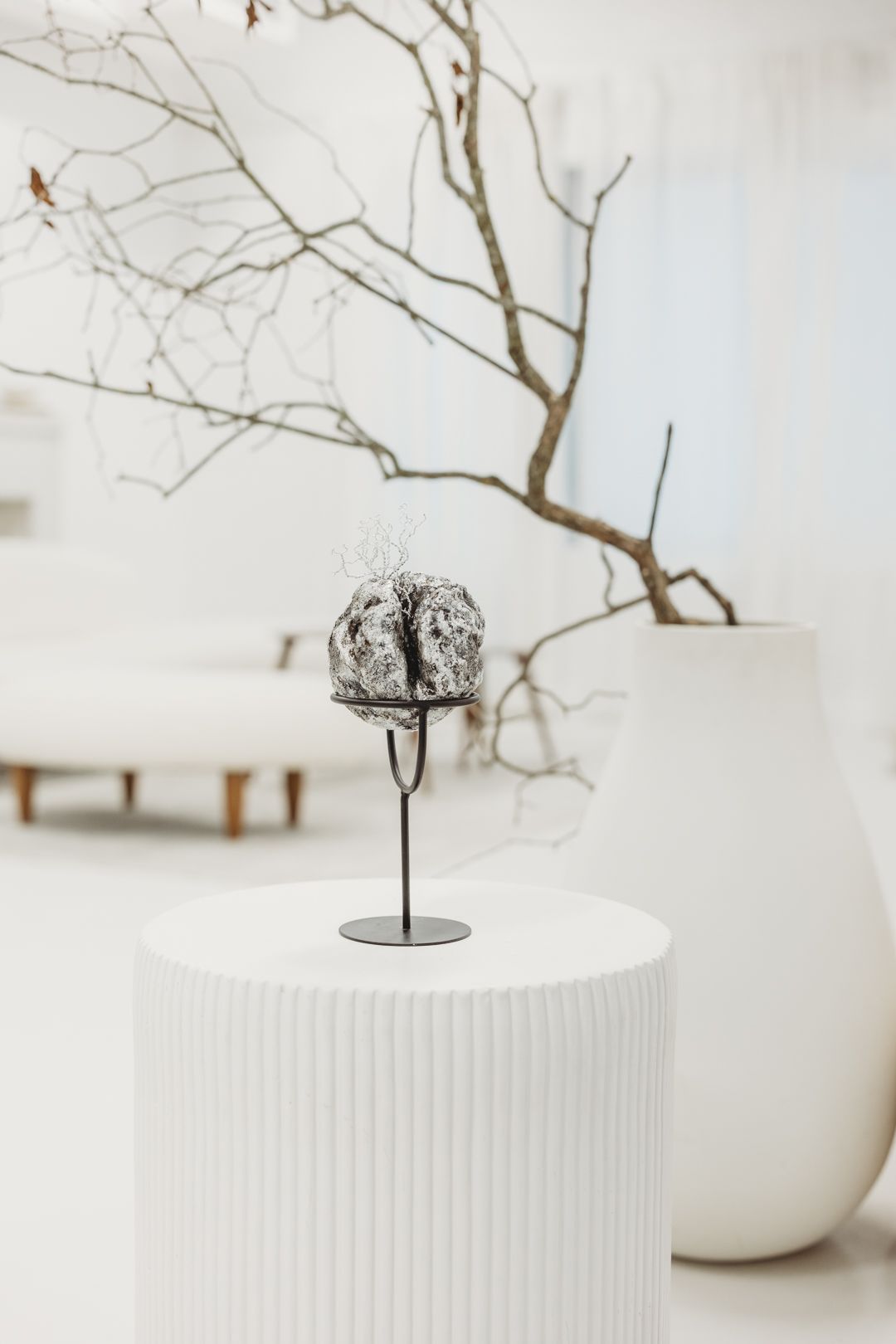 A textured stone sphere on a metal stand displayed on a white pedestal in a minimalist gallery with bare branches.