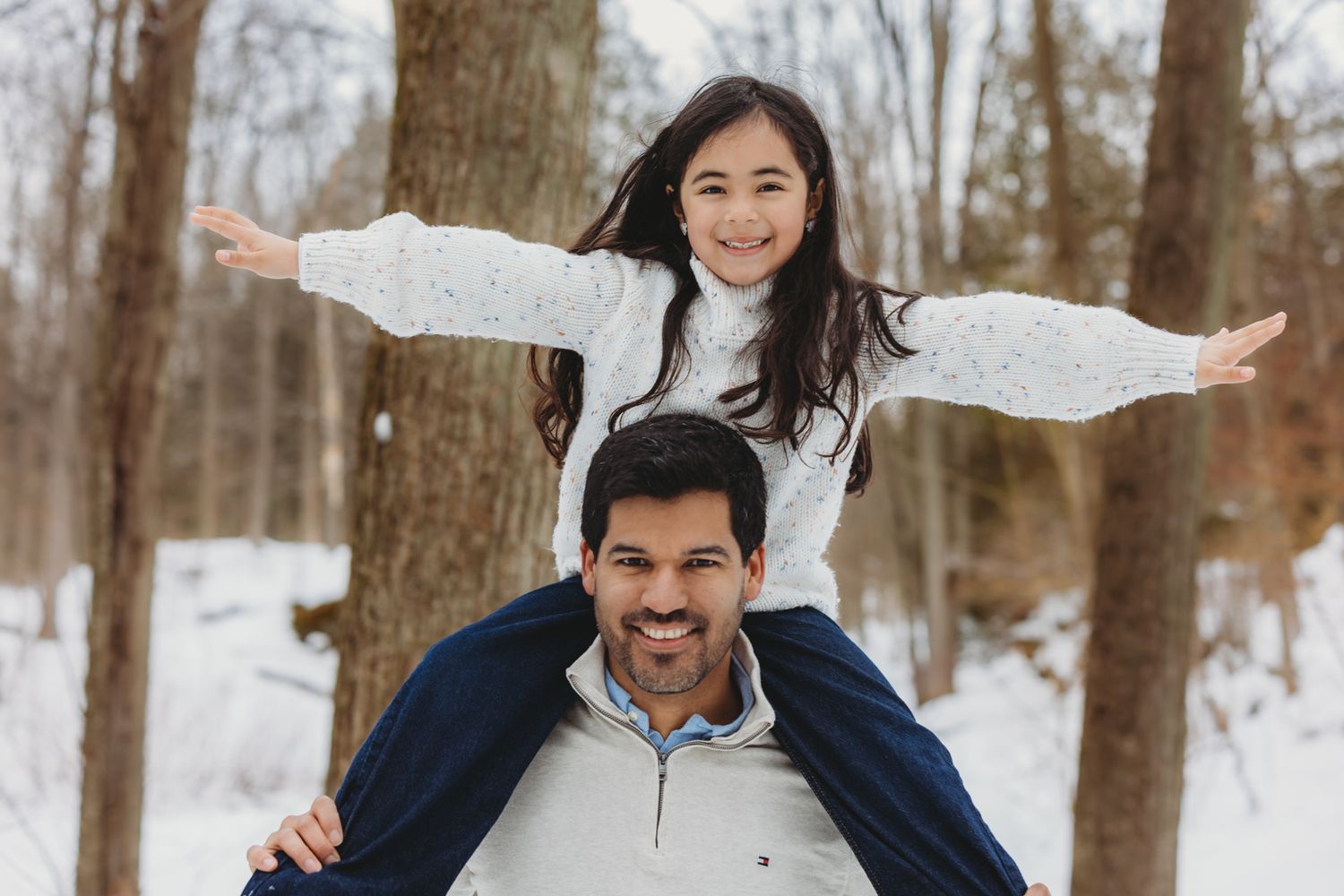 A smiling girl spreads her arms wide while sitting on her father's shoulders in a snowy winter forest.