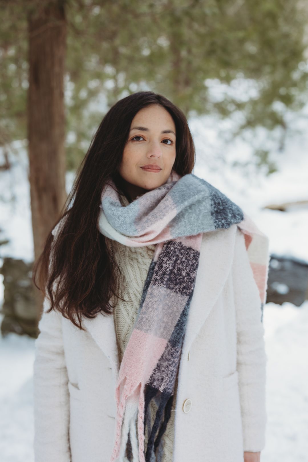 Young woman in white coat and plaid scarf standing in snowy winter forest.