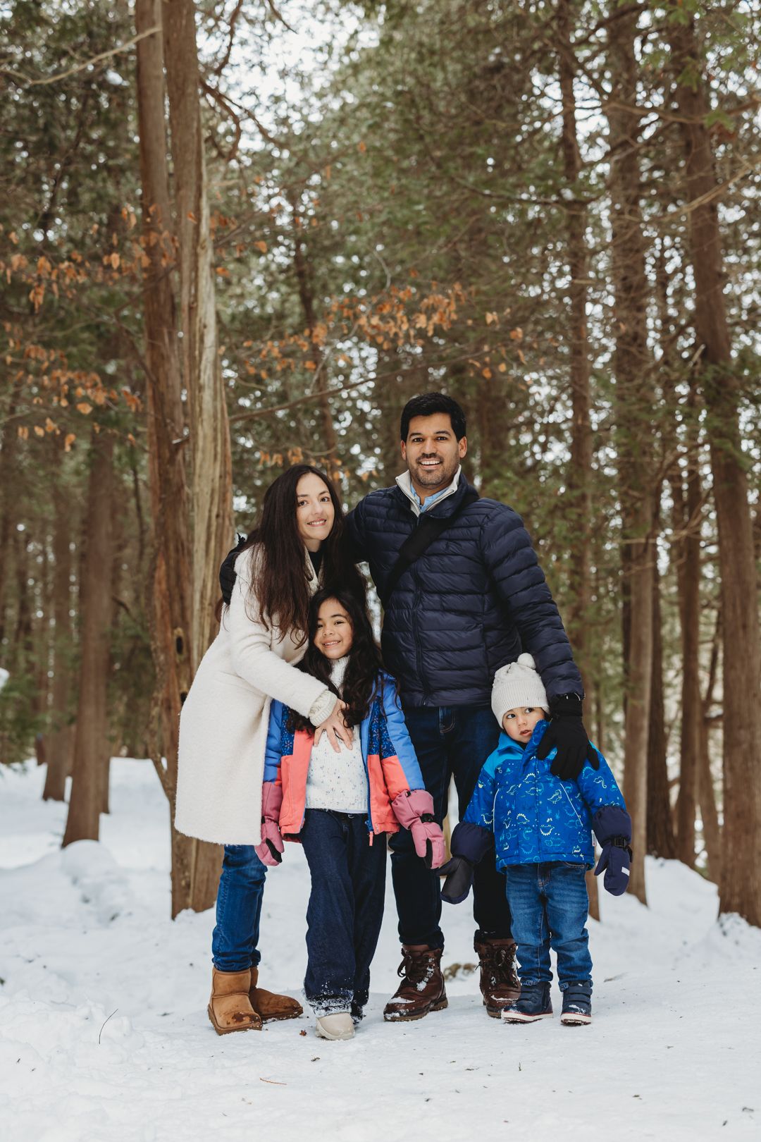 A happy family of four poses together in a snowy winter forest wearing colorful cold weather clothing.