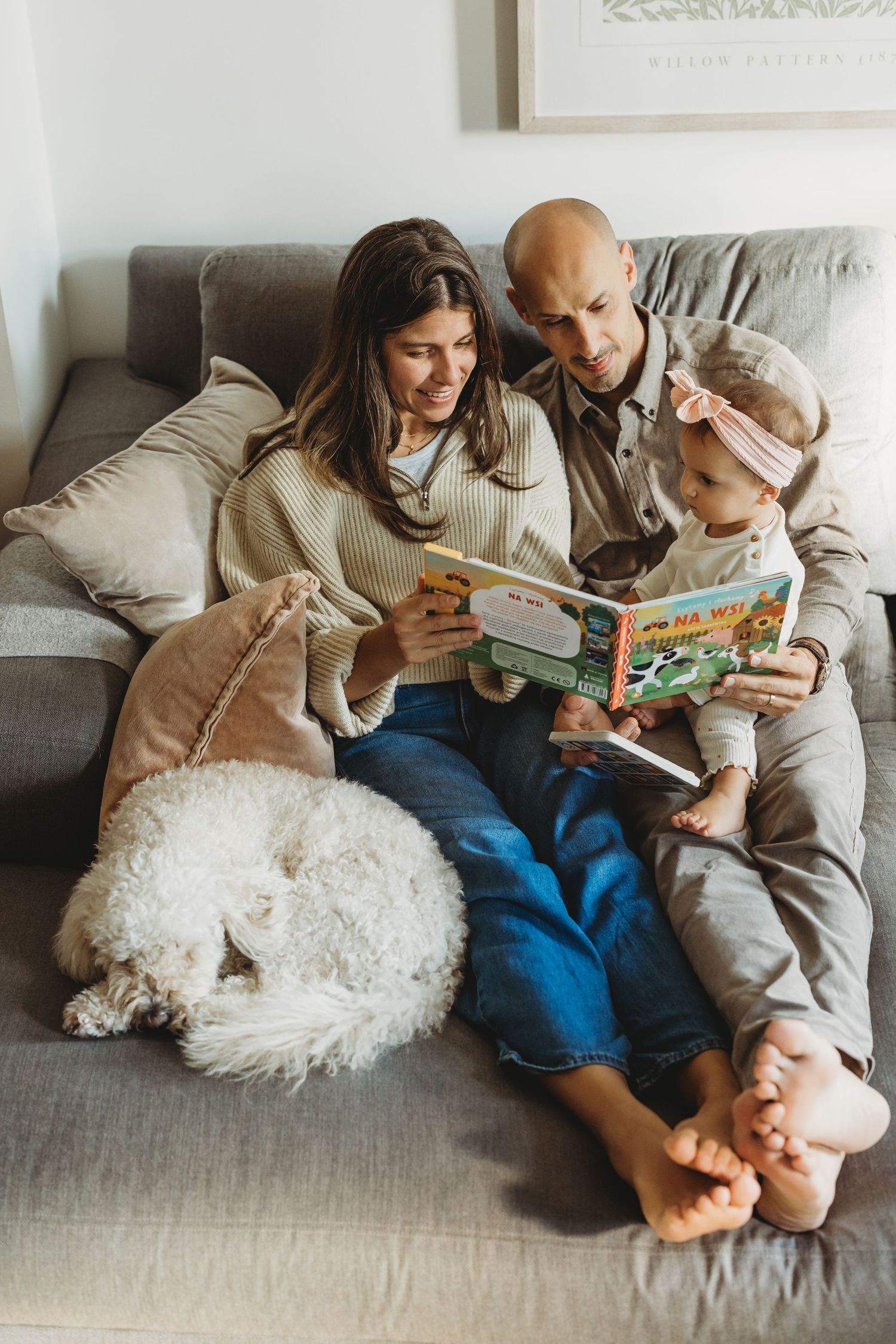 Family of three reading a colorful book together on a cozy sofa with a fluffy white dog resting nearby.