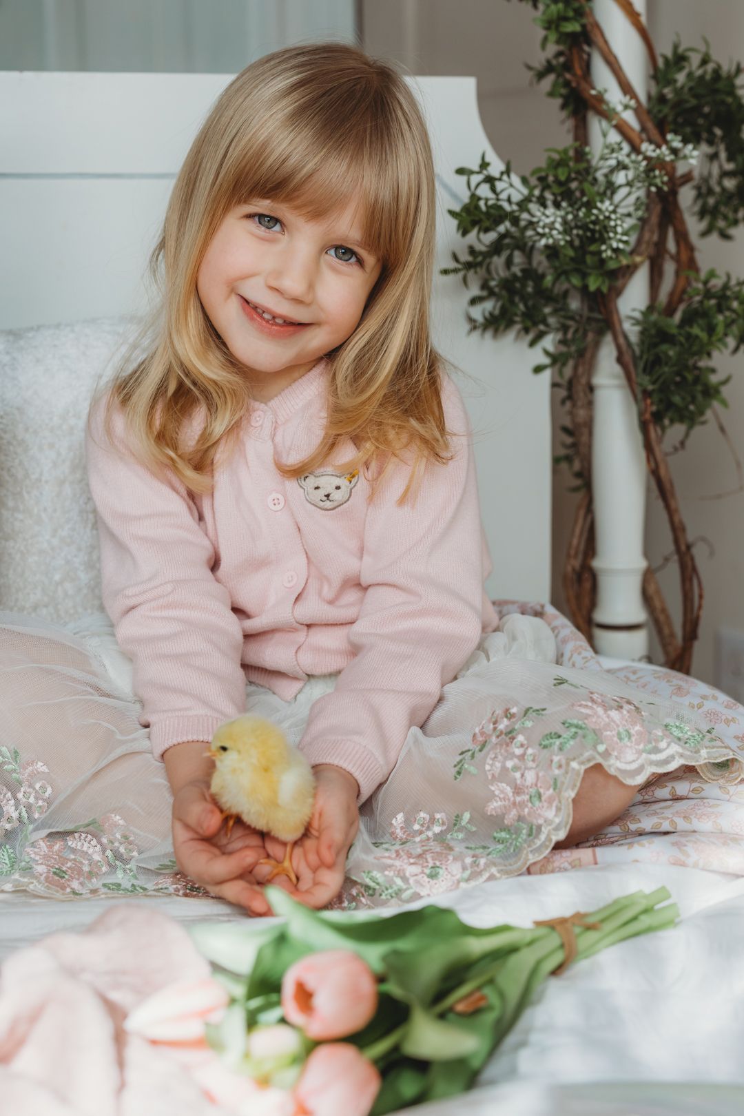 Smiling blonde girl in pink cardigan holds yellow baby chick on white bed with tulips nearby.