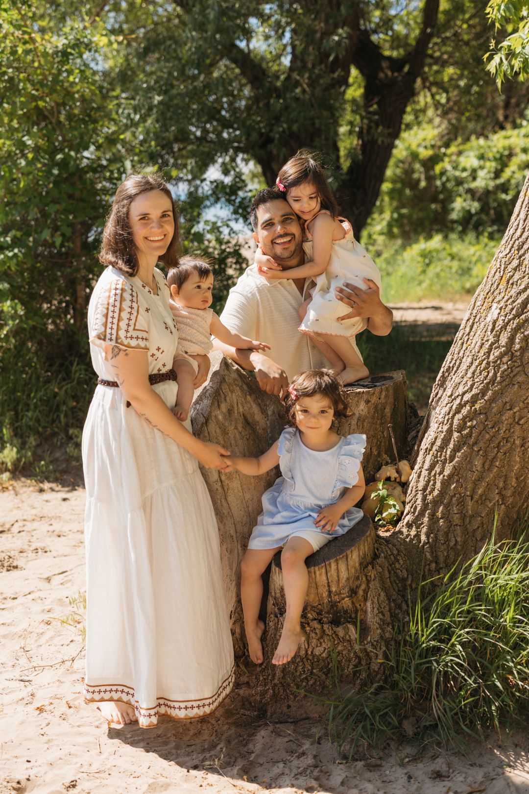Happy family with three young children posing by a tree stump in a sunny green park setting in Burlington, Ontario.