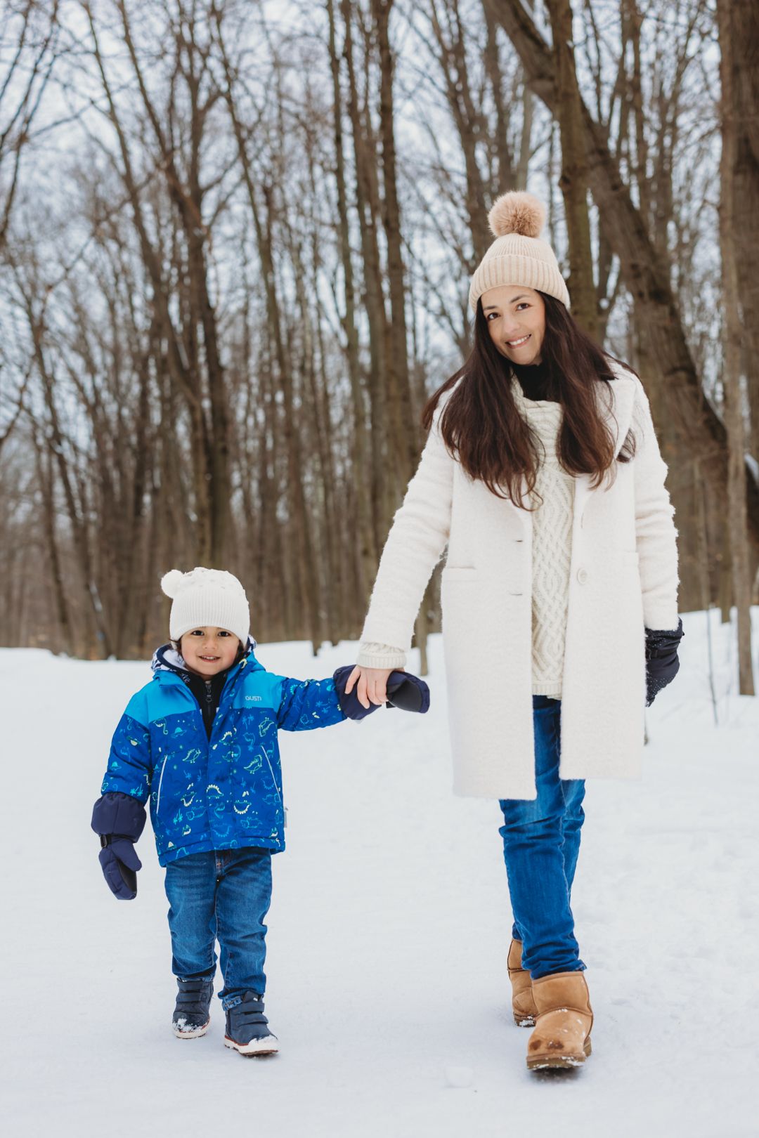 A smiling mother and young child walk hand-in-hand through a snowy winter park, both wearing cozy hats and winter coats.
