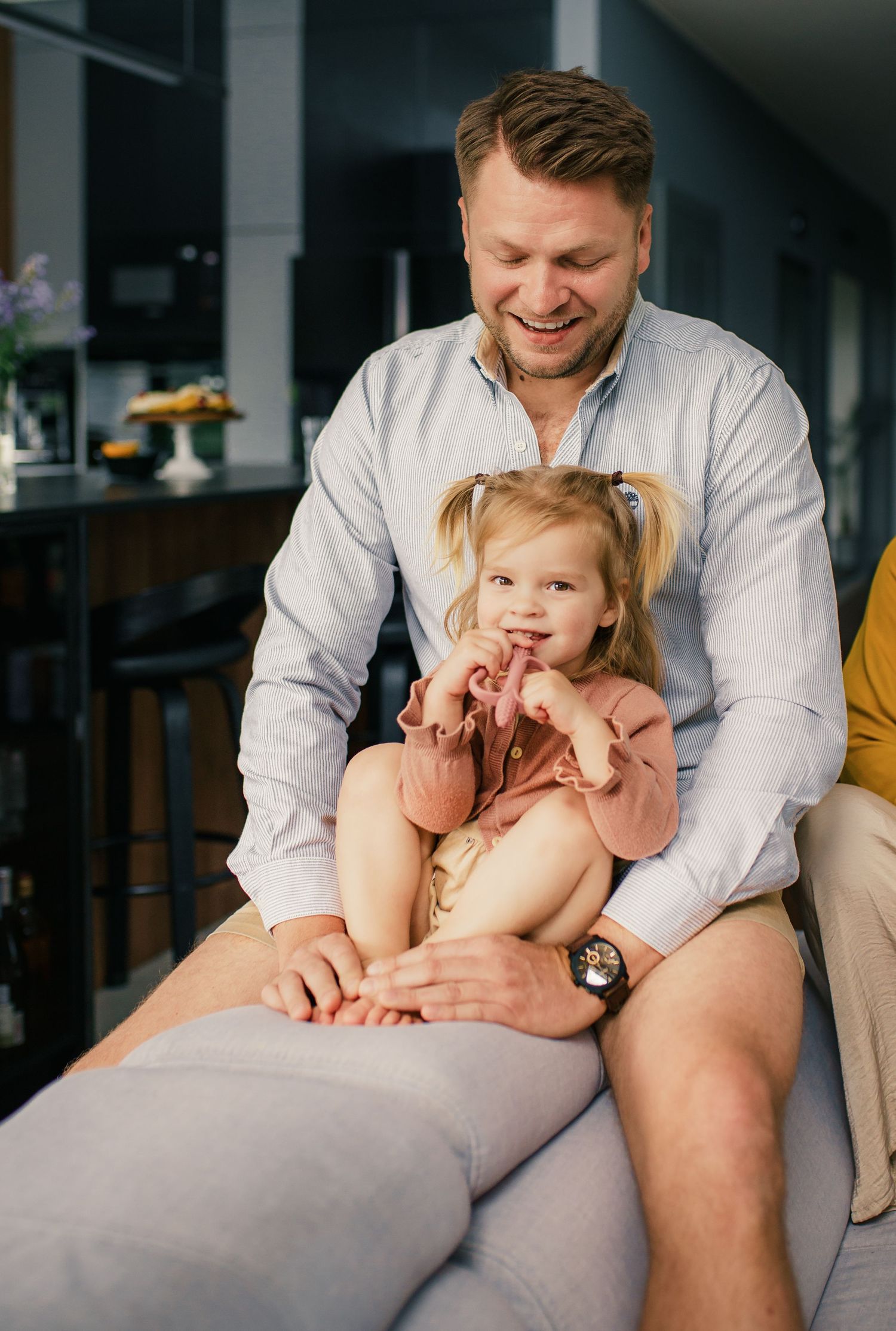 A cheerful father in a light blue shirt sits with his young daughter on a gray sofa at home.