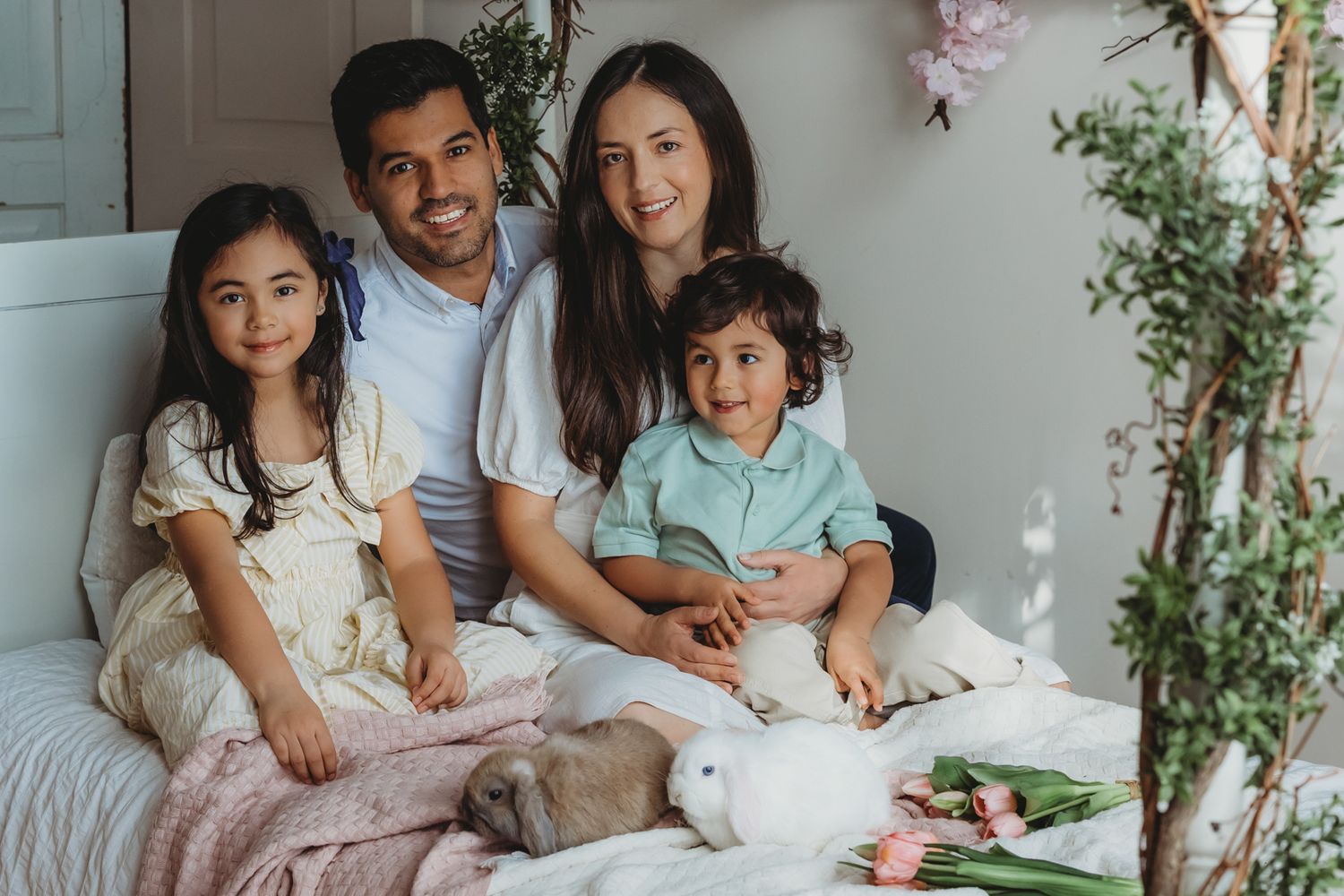 Happy family of four sitting on bed with white rabbits and pink tulips in spring setting.
