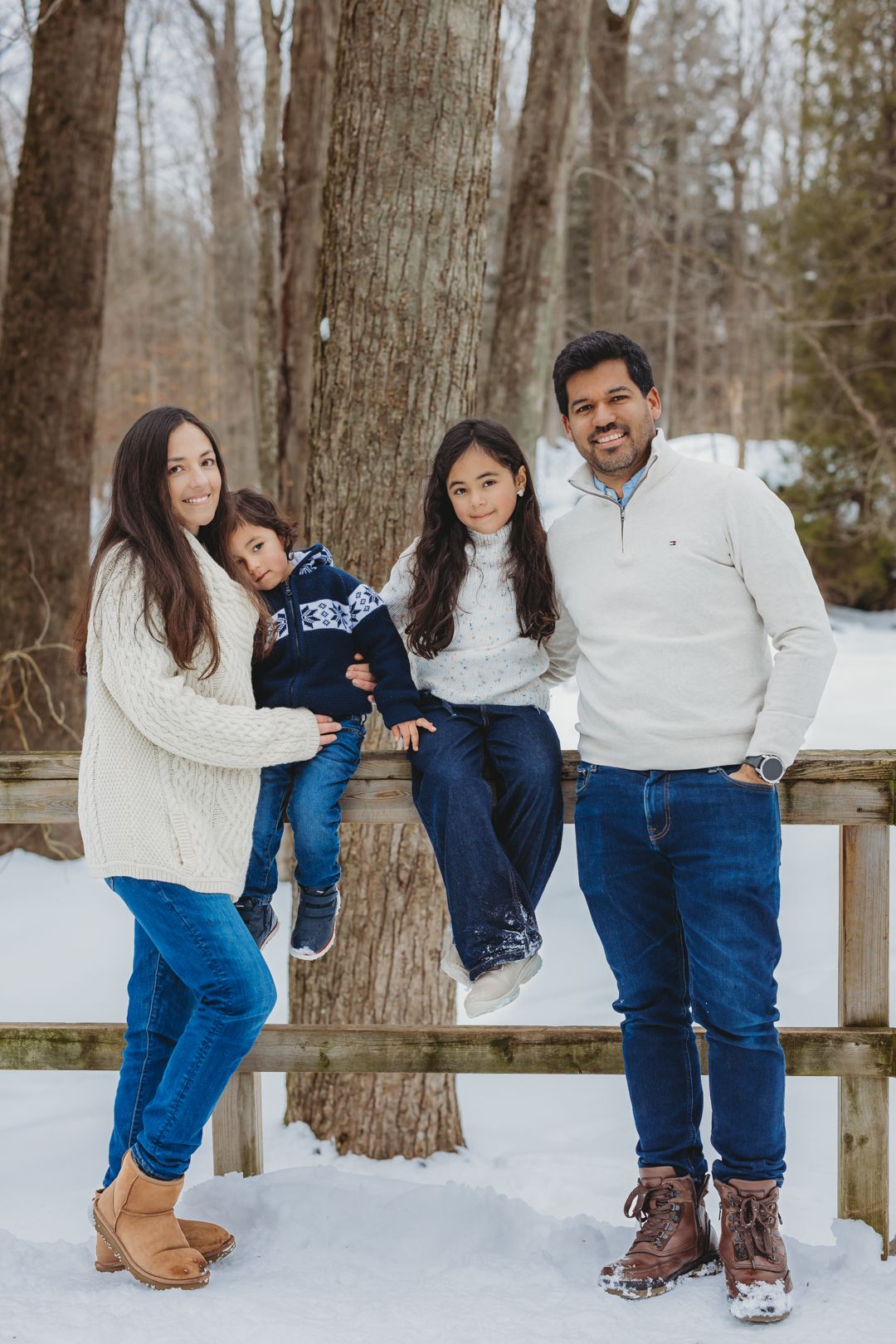 Family of four poses on snowy wooden bridge in matching white sweaters and jeans.