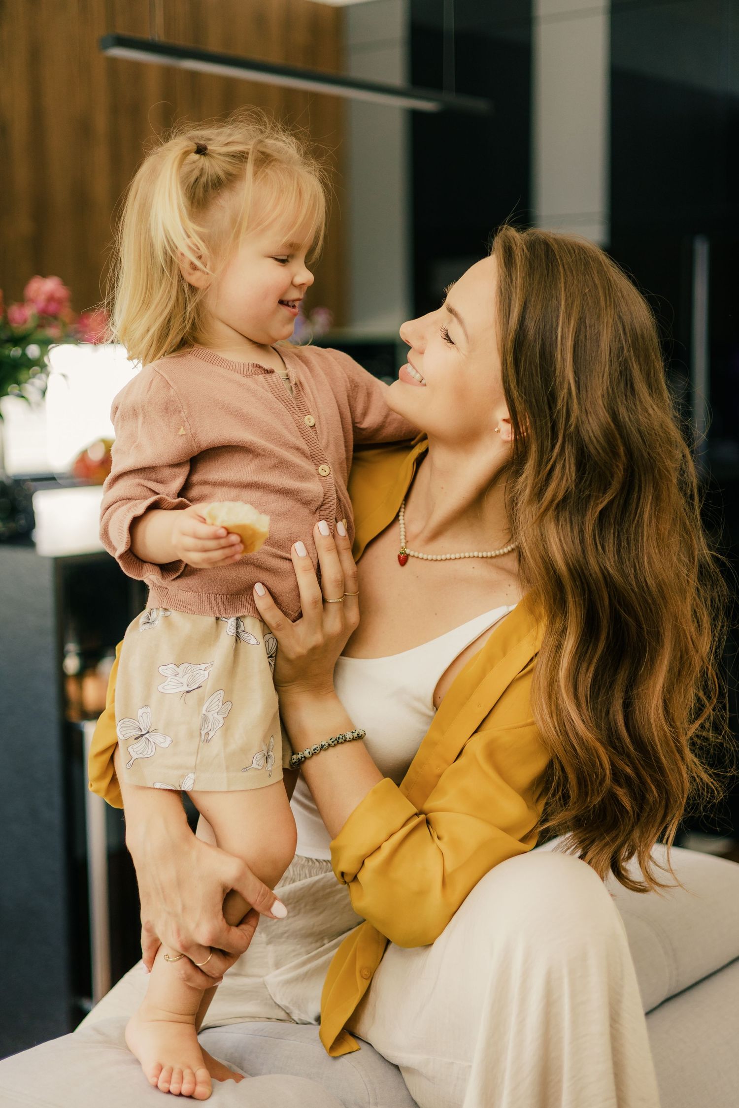 A loving mother in a yellow jacket lifts her smiling toddler daughter indoors in a bright modern home.
